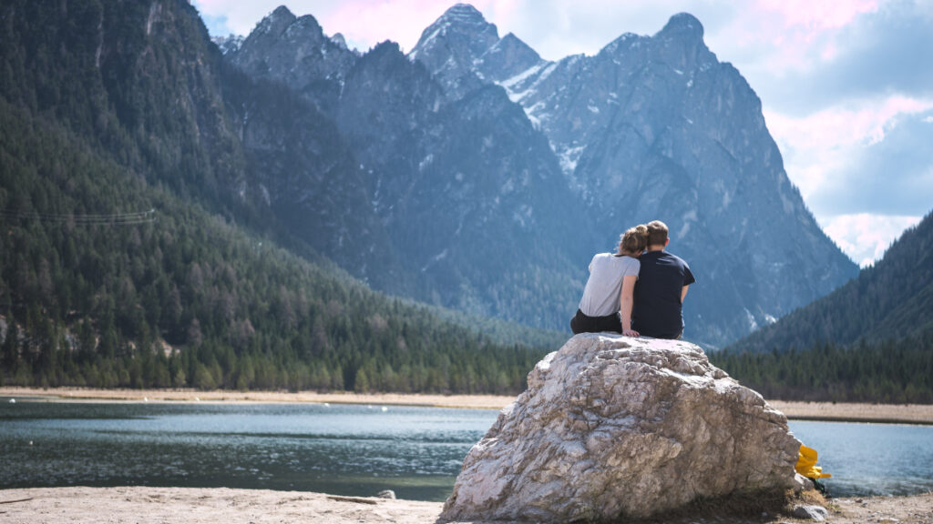 man and woman sitting on rock near body of water and mountain during daytime