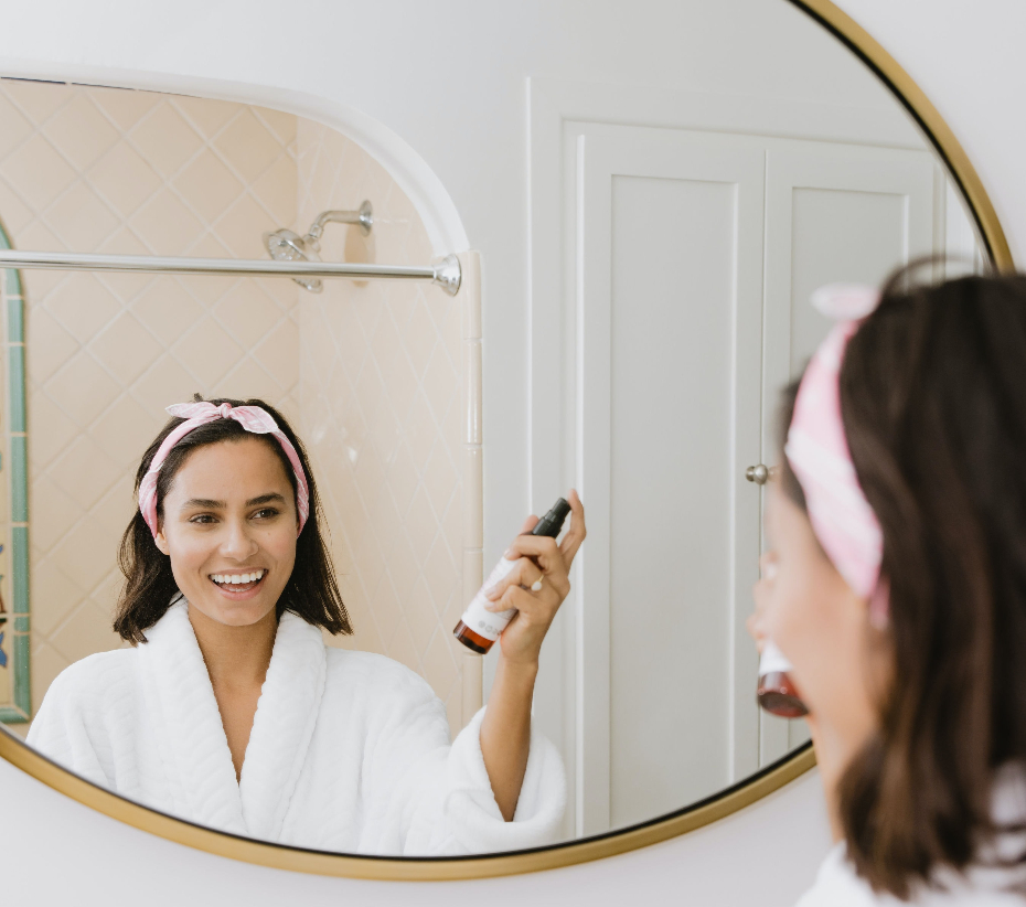 girl stands in front of mirror in a robe spraying on part of her skincare routine