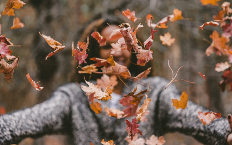 a girl wearing a sweater throws fall leaves up in the air while laughing and smiling