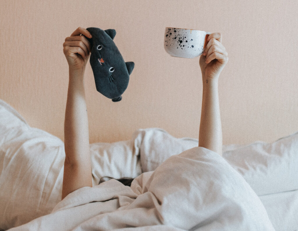 girl in white bedding holds up sleep mask and coffee mug