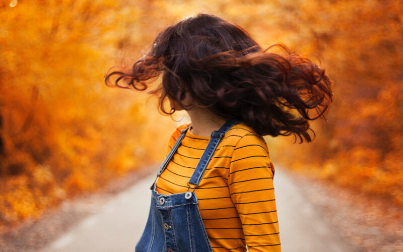 brunette girl wearing overalls and orange striped long-sleeve shirt looks back at fall leaves in the middle of the street