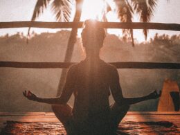 girl meditates on wooden balcony in front of a palm tree during a wellness experience